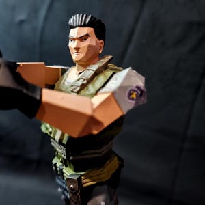 Może przedstawiać: Figurka akcji w stylu low-poly przedstawiająca postać z krótkimi ciemnymi włosami, ubrana w zieloną kamizelkę taktyczną na jasnej koszuli, ciemne spodnie i czarne rękawiczki. Figurka posiada naszywkę na ramieniu z okrągłym emblematem.