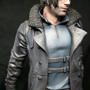 May include: A detailed figurine of a man with short, light brown hair and a serious expression. He wears a gray coat with a faux fur collar, a light blue shirt, and black gloves. The coat has multiple buttons and straps.