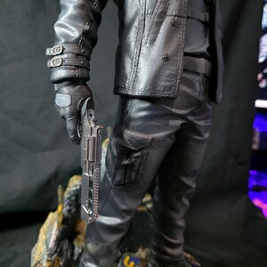 May include: A detailed figurine of a figure in a black leather jacket, pants, and boots, holding a silver handgun. The figure is standing on a rocky base with gold and blue accents. The jacket has button details and the pants have a pocket.