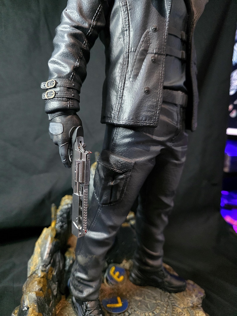 May include: A detailed figurine of a figure in a black leather jacket, pants, and boots, holding a silver handgun. The figure is standing on a rocky base with gold and blue accents. The jacket has button details and the pants have a pocket.