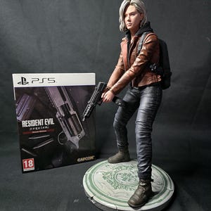 以下が含まれることがあります： Resident Evilのキャラクターの精巧なフィギュア。銃を持ち、茶色の革のジャケット、黒いパンツ、バックパックを着用。フィギュアは丸い台座に立っています。背景にはPS5のゲームボックスが見えます。