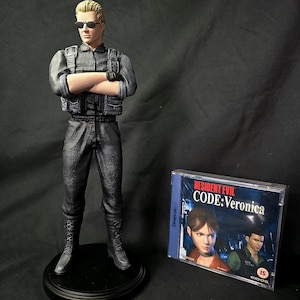 Könnte beinhalten: Detaillierte Figur von Albert Wesker, einer Figur aus der Resident Evil-Videospielserie, mit Sonnenbrille und taktischer Weste. Die Figur steht auf einem schwarzen Sockel. Daneben eine Kopie von Resident Evil Code: Veronica für Dreamcast.