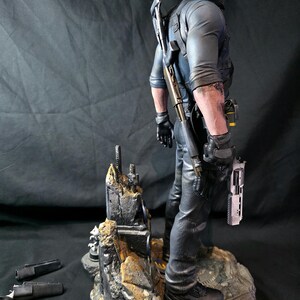 May include: A detailed figurine of a man in tactical gear, holding a pistol. He stands on a rocky base with additional weapons and accessories scattered around. The figure is dressed in black pants, a blue shirt, and black gloves.