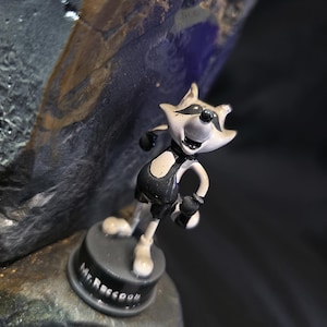 May include: A small, cartoonish figurine of a raccoon in a black and white outfit, standing on a black base. The base is inscribed with "Mr. Raccoon" in white lettering. The figurine is set against a dark, textured background.