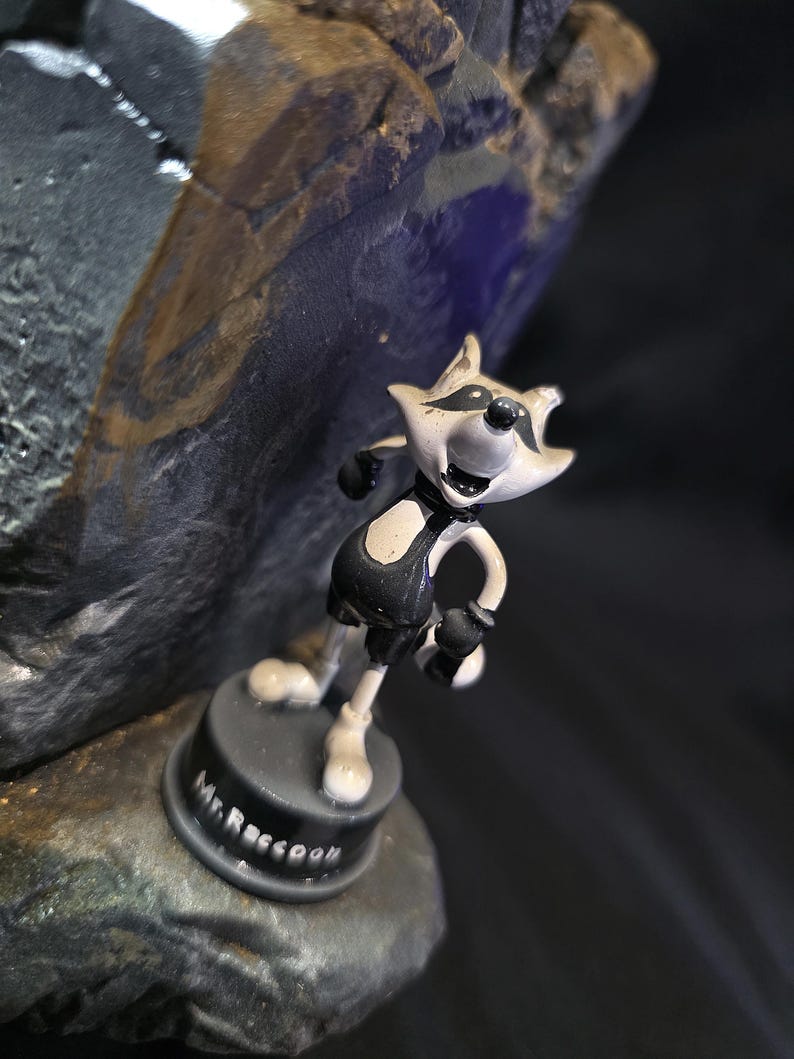 May include: A small, cartoonish figurine of a raccoon in a black and white outfit, standing on a black base. The base is inscribed with "Mr. Raccoon" in white lettering. The figurine is set against a dark, textured background.