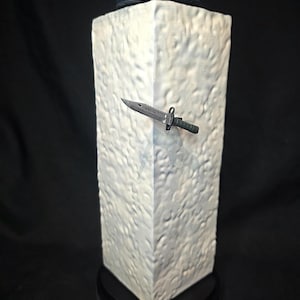 May include: A white textured rectangular block featuring a dark gray combat knife embedded in the side. A dark blue beret with a circular emblem sits on top. The base is black with the text TOFU printed in white letters on the front edge.