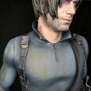 May include: A detailed figurine of a man with short, styled gray hair and blue eyes. He wears a blue-gray tactical shirt with a zipper and black straps. The figure has a serious expression and is set against a dark background.