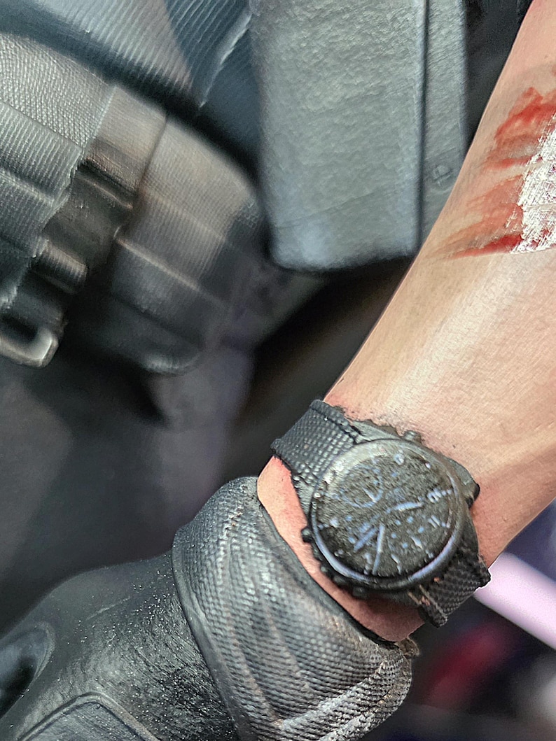 May include: Close-up of a black wristwatch with a textured face and a black fabric band. The watch is on a wrist with a visible wound. The watch face has multiple dials and hands.