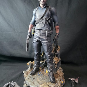 May include: A detailed figurine of a character in a blue shirt, black pants, and boots, holding a gun. The figure stands on a rocky base with a metallic emblem. Additional weapons are placed near the base. The figure has a serious expression.