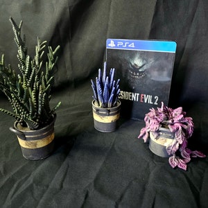 May include: Three miniature potted plants and a PS4 game case. The plants are green, purple, and blue, in black and gold-banded pots. The game case features "Resident Evil 2" and a monster graphic.