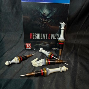 May include: Four chess pieces, a queen, a knight, a rook, and a pawn, have been modified to look like weapons. The pieces are painted black, white, gold, and red. The pieces are lying on a black surface in front of a video game case for Resident Evil 2. The case is blue and has the PlayStation 4 logo on it.