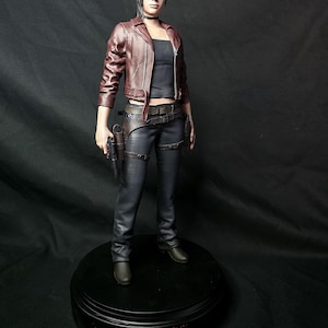 Può includere: Una action figure da collezione di un personaggio con capelli scuri, che indossa una giacca in pelle marrone scuro, un top nero e pantaloni scuri con una fondina. La figura poggia su una base nera rotonda con la scritta Resident Evil in rosso.