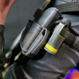 May include: Close-up of tactical gear, featuring a black pouch and a cylindrical object with a yellow band. The gear is attached to a dark-colored garment, with visible straps and stitching. The overall aesthetic suggests a military or tactical theme.