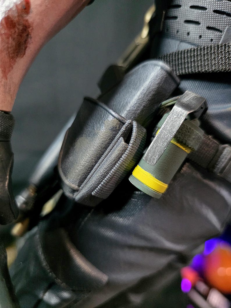 May include: Close-up of tactical gear, featuring a black pouch and a cylindrical object with a yellow band. The gear is attached to a dark-colored garment, with visible straps and stitching. The overall aesthetic suggests a military or tactical theme.