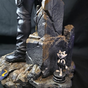 May include: A detailed figurine featuring a character in black pants and boots, standing on a rocky base. A small cartoon raccoon figure labeled "Mr. Raccoon" is also present. The base has a blue and yellow emblem.