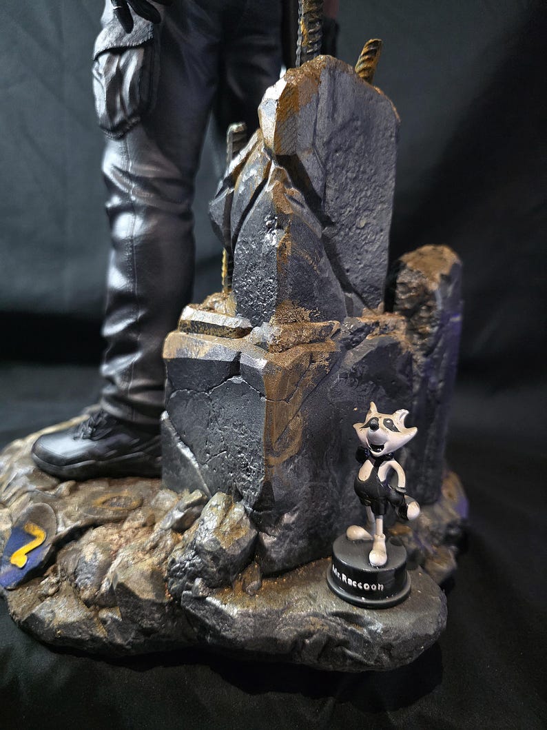 May include: A detailed figurine featuring a character in black pants and boots, standing on a rocky base. A small cartoon raccoon figure labeled "Mr. Raccoon" is also present. The base has a blue and yellow emblem.