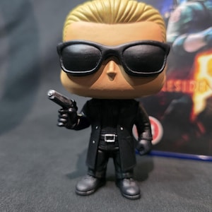 May include: A vinyl figure of a character from the Resident Evil video game series. The figure has a large head, black sunglasses, and a black outfit. It is holding a silver handgun. The figure is approximately 4 inches tall.