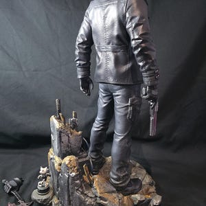 May include: A black figurine of a person in a leather jacket and pants, holding a gun. The figure stands on a rocky base with additional small figurines and a weapon. The overall color scheme is dark, with detailed textures.