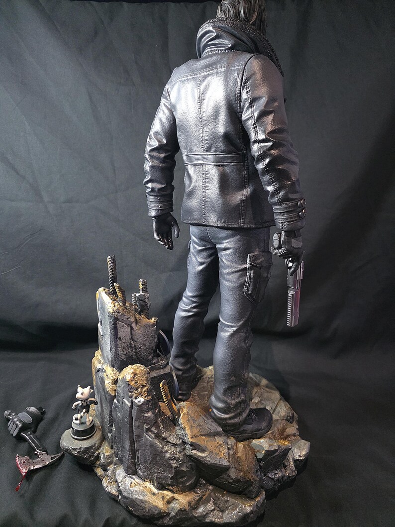 May include: A black figurine of a person in a leather jacket and pants, holding a gun. The figure stands on a rocky base with additional small figurines and a weapon. The overall color scheme is dark, with detailed textures.
