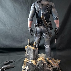 May include: A detailed figurine of a character in tactical gear, holding a pistol and a shotgun. The figure stands on a rocky base with additional weapons and a small figurine. The figure is dressed in black and gray clothing.