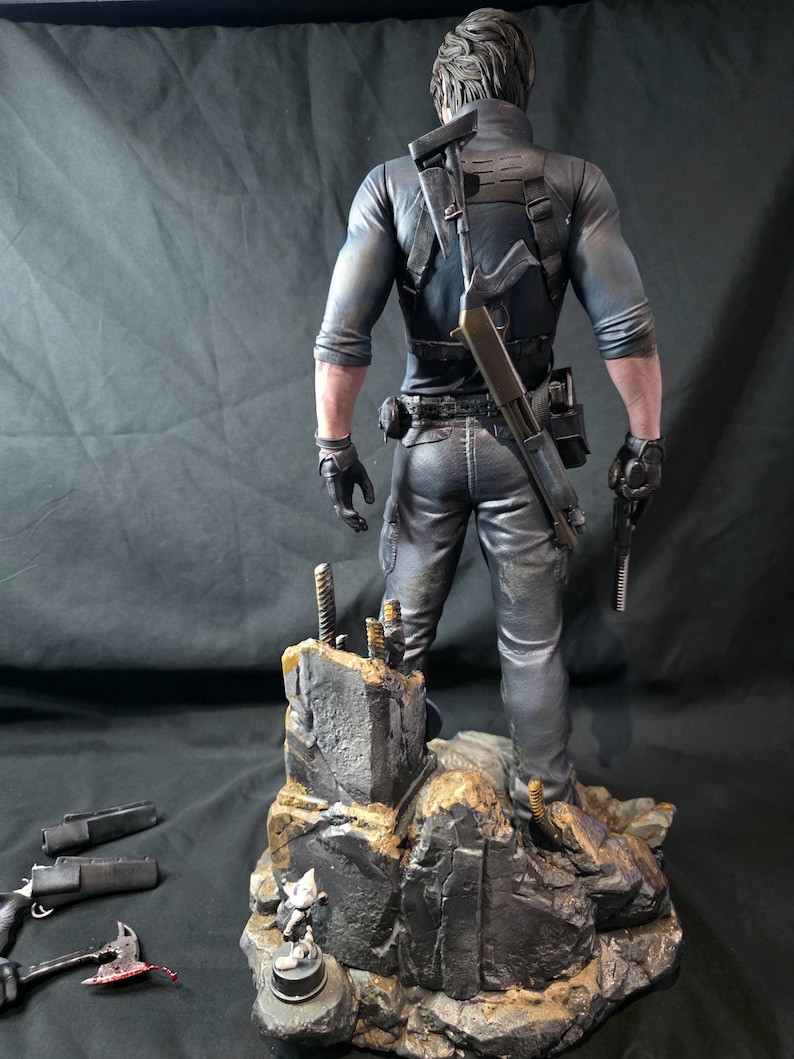 May include: A detailed figurine of a character in tactical gear, holding a pistol and a shotgun. The figure stands on a rocky base with additional weapons and a small figurine. The figure is dressed in black and gray clothing.