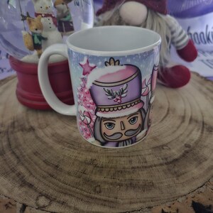 May include: A white ceramic mug with a colourful design featuring a nutcracker in a purple and pink hat. The mug has a blue and white winter scene background with a candy cane and a Christmas tree.