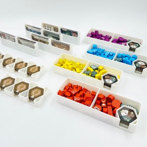 May include: White plastic organisers for board game pieces. The organisers have multiple compartments for storing different coloured game pieces, cards, and tokens.