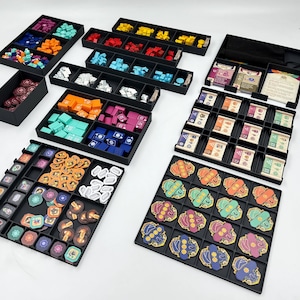 May include: A collection of black game organizers filled with colorful game pieces, cards, and tokens. The pieces are in various shapes and colors, including blue, orange, green, and purple. The organizers are arranged on a white surface.