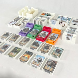 Könnte beinhalten: Eine Sammlung weißer Spielzeug-Organizer aus Kunststoff mit verschiedenfarbigen Kartenhaltern. Die Organizer halten Spielkarten und Spielsteine. Die Karten zeigen Illustrationen von Fantasy-Charakteren und -Szenen. Die Organizer sind auf einer weißen Oberfläche angeordnet.