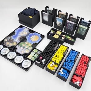 May include: A black organiser for a board game with multiple compartments for storing game pieces, cards, and dice. The organiser includes a variety of compartments for different sized pieces, including small compartments for dice and larger compartments for cards and game boards.