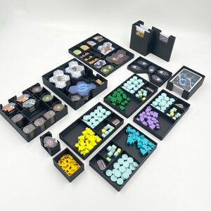 May include: A set of black plastic organisers for board game pieces. The organisers are filled with various coloured plastic pieces, including blue, green, yellow, and purple. The organisers are designed to fit into a larger box, and they are perfect for storing and organising board game components.