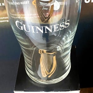 May include: A clear Guinness pint glass with the Guinness logo and harp design. The glass has the word "Guinness" in white lettering. The glass is on a black surface.