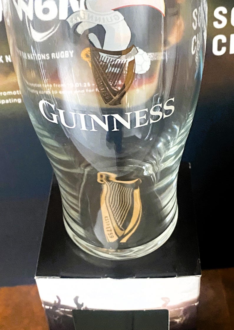 May include: A clear Guinness pint glass with the Guinness logo and harp design. The glass has the word "Guinness" in white lettering. The glass is on a black surface.