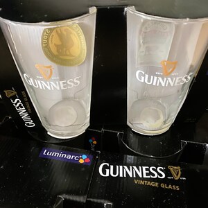 May include: Two clear Guinness pint glasses with the Guinness logo and harp emblem are displayed. The glasses are in a black box with the words "Guinness Vintage Glass" printed in white. The box also features the Luminarc logo.