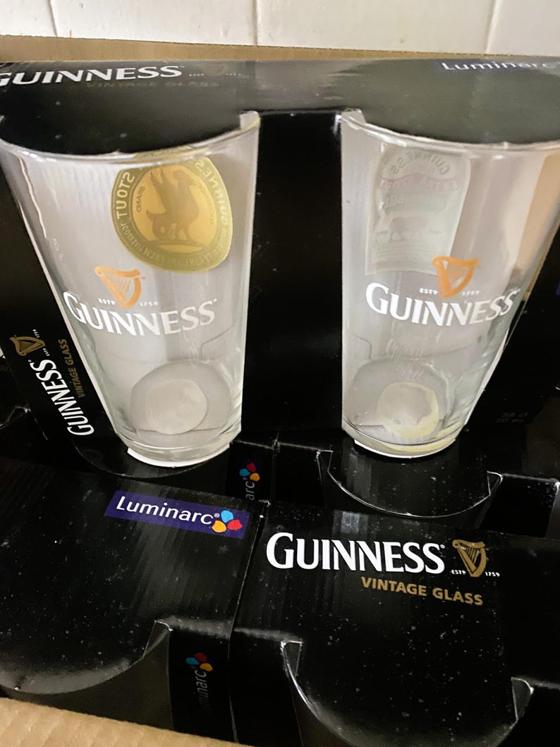 May include: Two clear Guinness pint glasses with the Guinness logo and harp emblem are displayed. The glasses are in a black box with the words "Guinness Vintage Glass" printed in white. The box also features the Luminarc logo.