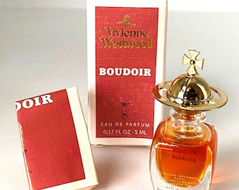 May include: A small, clear glass bottle of Vivienne Westwood Boudoir Eau de Parfum. The bottle has a gold orb stopper with a cross on top. The bottle is sitting on a white surface. The box for the perfume is also visible in the image. The box is white with gold lettering and a red background. The box says "Vivienne Westwood" and "Boudoir". The box also says "Eau de Parfum" and "0.17 fl oz - 5 ml".