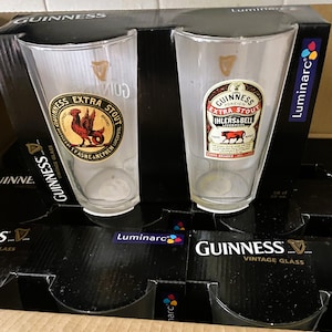 May include: Two clear Guinness pint glasses with vintage labels featuring a rooster and a bull. The glasses are in a black box labeled "Guinness Vintage Glass" and "Luminarc". The glasses are empty and ready to be filled.