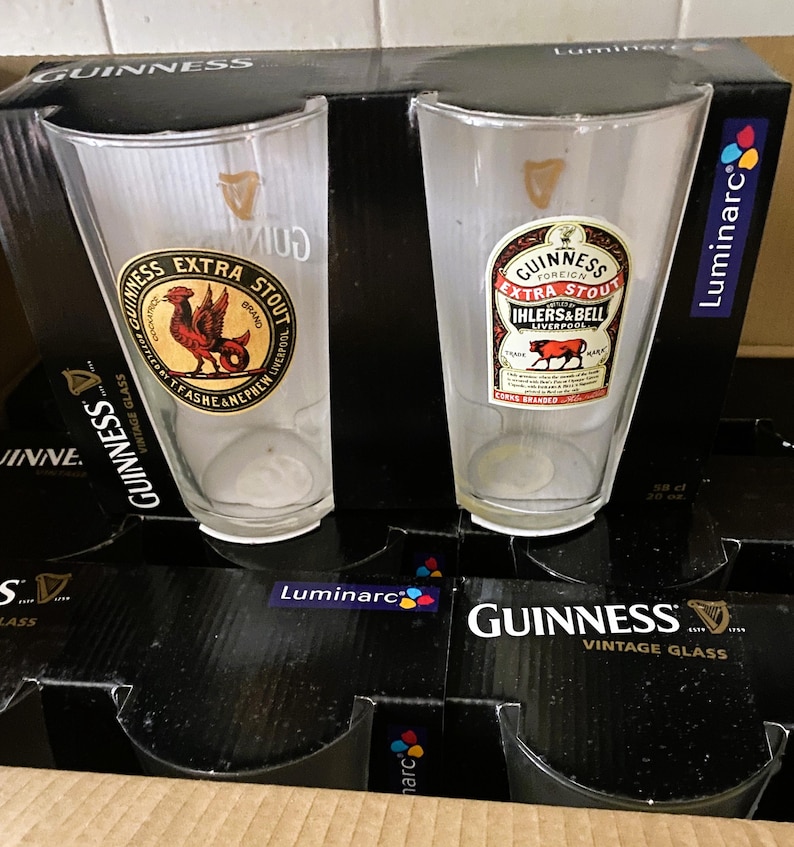 May include: Two clear Guinness pint glasses with vintage labels featuring a rooster and a bull. The glasses are in a black box labeled "Guinness Vintage Glass" and "Luminarc". The glasses are empty and ready to be filled.