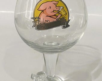 May include: Clear glass beer glass with a stem featuring a small pig figurine. The glass has a large bowl with a pig illustration holding a beer mug. The illustration is inside a yellow circle with the text "Rince Co." on a banner.