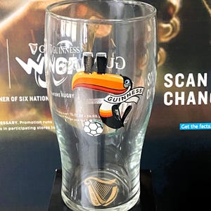 May include: A clear Guinness pint glass with a toucan logo, two black pints, and a soccer ball. The glass has the Guinness logo and a harp emblem. The background features promotional material for Guinness and rugby.