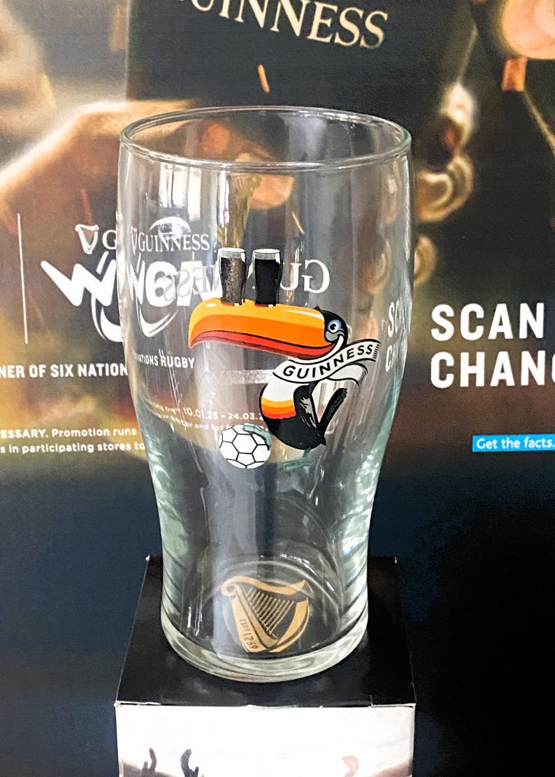 May include: A clear Guinness pint glass with a toucan logo, two black pints, and a soccer ball. The glass has the Guinness logo and a harp emblem. The background features promotional material for Guinness and rugby.