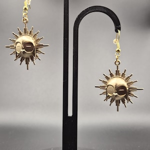 Könnte beinhalten: Ein Paar goldfarbene, sonnenförmige Ohrringe. Jeder Ohrring zeigt ein Sonnengesicht mit Strahlen. Die Ohrringe sind an goldfarbenen Ohrhaken befestigt. Die Ohrringe werden auf einem schwarzen Ständer vor grauem Hintergrund präsentiert.