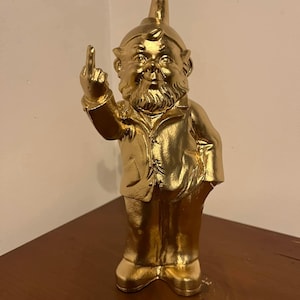 May include: A golden garden gnome statue with a beard, hat, and jacket, giving the middle finger. The gnome is standing on a wooden surface. The statue is a humorous and unique garden decoration.