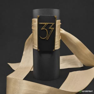 May include: A matte black cylindrical container wrapped with a wide, champagne-colored satin ribbon. A rectangular black plaque is centered on the front, featuring the gold-colored number 33 in a stylized font with decorative corner accents.