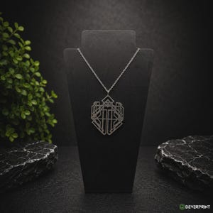 Può includere: Una collana color argento con un ciondolo geometrico traforato dal design simmetrico e angolare. Il ciondolo è sospeso a una delicata catenina color argento e presenta un complesso motivo lineare che forma una sagoma simile a uno scudo.
