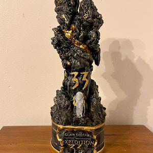 May include: A black and gold decorative sculpture with the text "33" and "Clair Obscur Expedition 33". The sculpture has a textured, rocky appearance with gold accents.