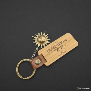 Pode incluir: Um porta-chaves com uma etiqueta retangular de bambu gravada com o texto EXPEDITION 33, presa a uma tira de couro castanho com um rebite de latão. Um pendente de sol em metal dourado com um rosto está ligado por uma corrente ao anel.