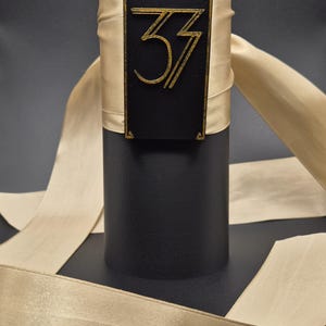 Peut inclure: Un objet cylindrique noir avec une étiquette rectangulaire dorée portant le chiffre "33" dans une police stylisée. L'objet est enveloppé d'un ruban beige. Le fond est sombre.