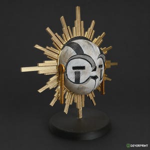 May include: A decorative sculpture featuring a central white mask with black markings, surrounded by a gold sunburst frame with radiating rays. The mask is mounted on a circular black base, and the entire piece displays a textured finish.