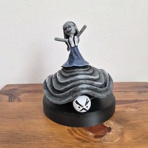 May include: A whimsical figurine featuring a stylized figure in a dress with arms outstretched, atop a layered, textured base. The base is black, and a small white ball with a black face is nestled beneath the figure. The figurine is set on a wooden surface.